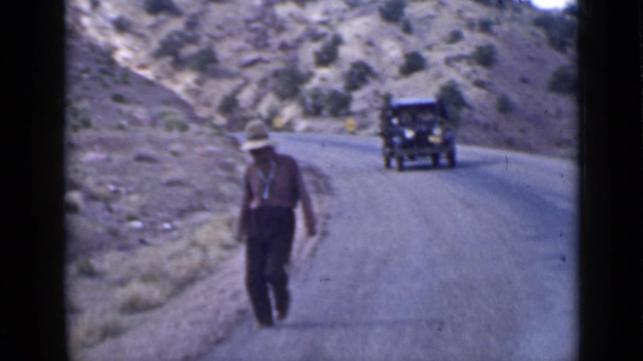 Man in a hat walking along a mountain road as a dark vintage car approaches.