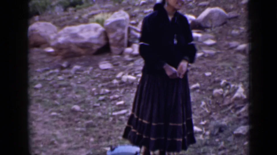 1939 Utah frame showing a woman in a long dark dress standing on rocky ground beside large stones in an arid landscape.