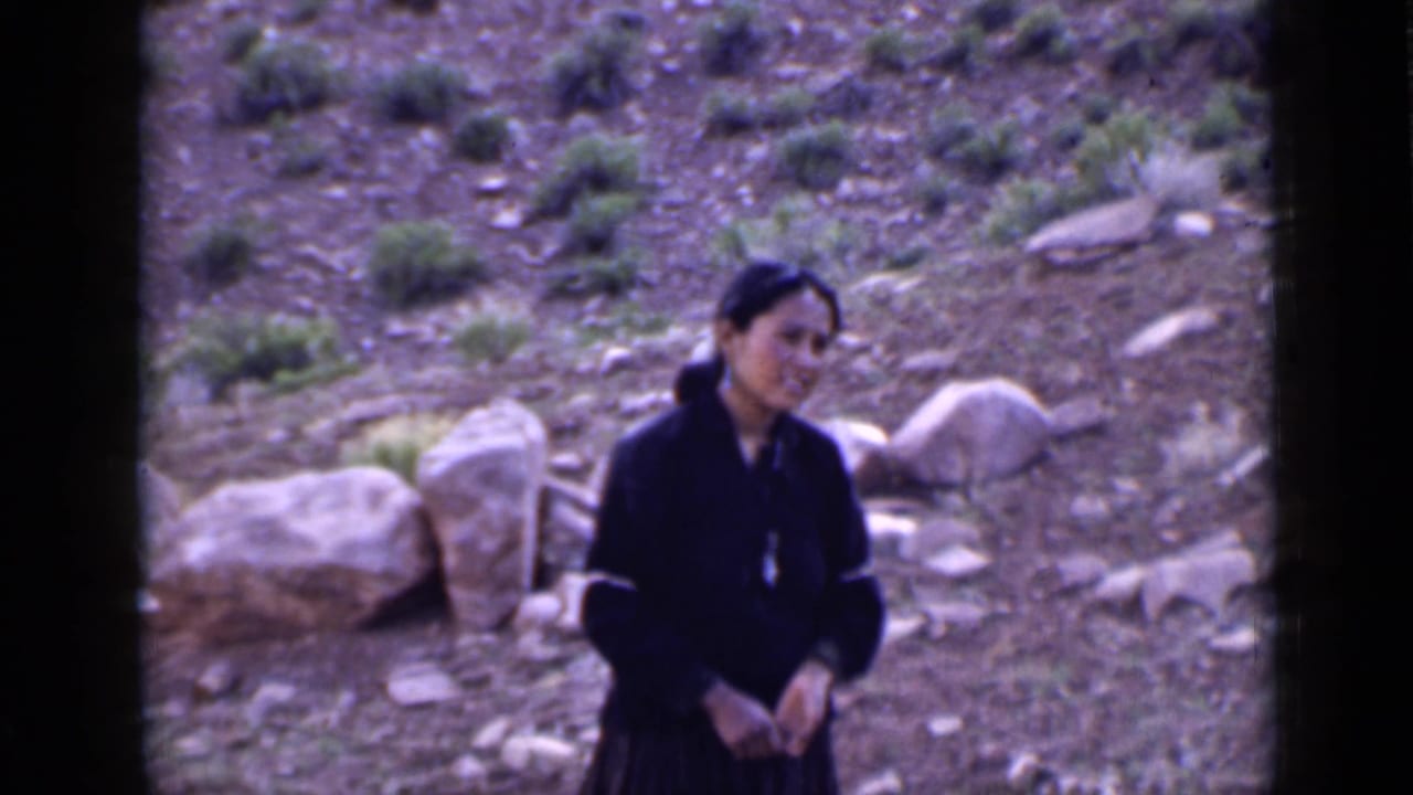 Woman in dark clothing standing on a rocky hillside with shrubs and large stones behind her.