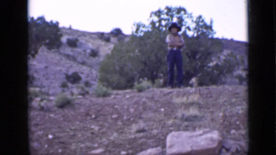 In 1939 Utah footage, a man wearing a cowboy hat stands with arms crossed on an arid hillside near small trees.