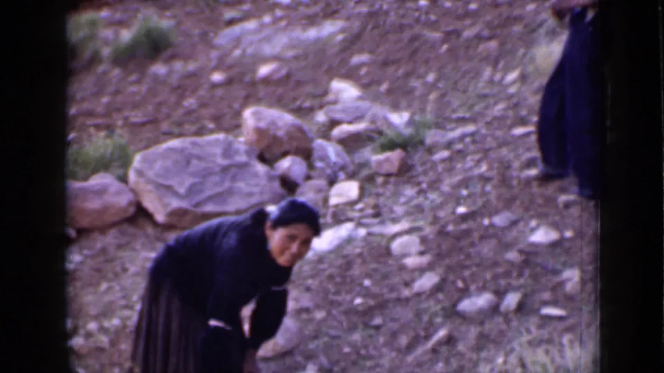A woman in dark clothing bends near rocks on a dry Utah hillside in archival 8mm footage.