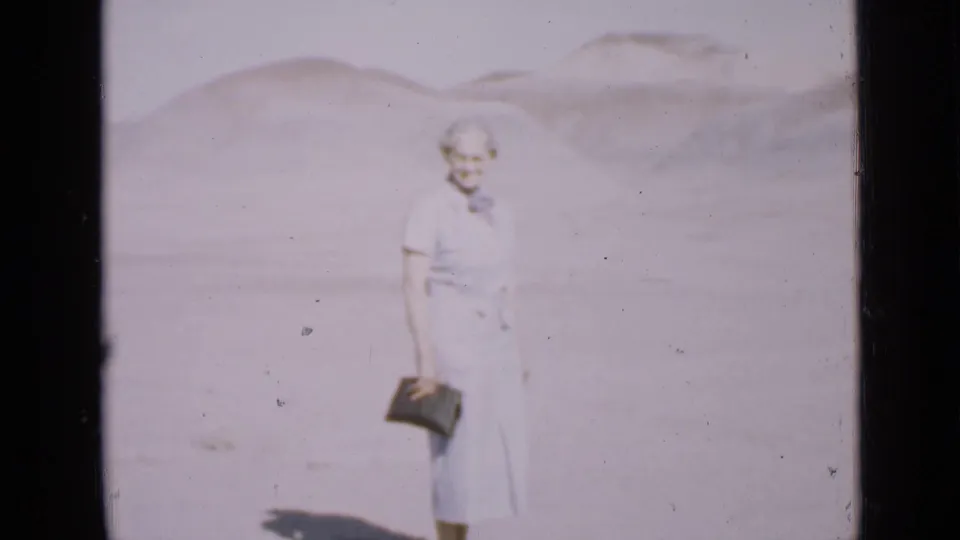 A woman in a light period dress stands in the Utah desert, holding a small dark bag with pale hills behind her.
