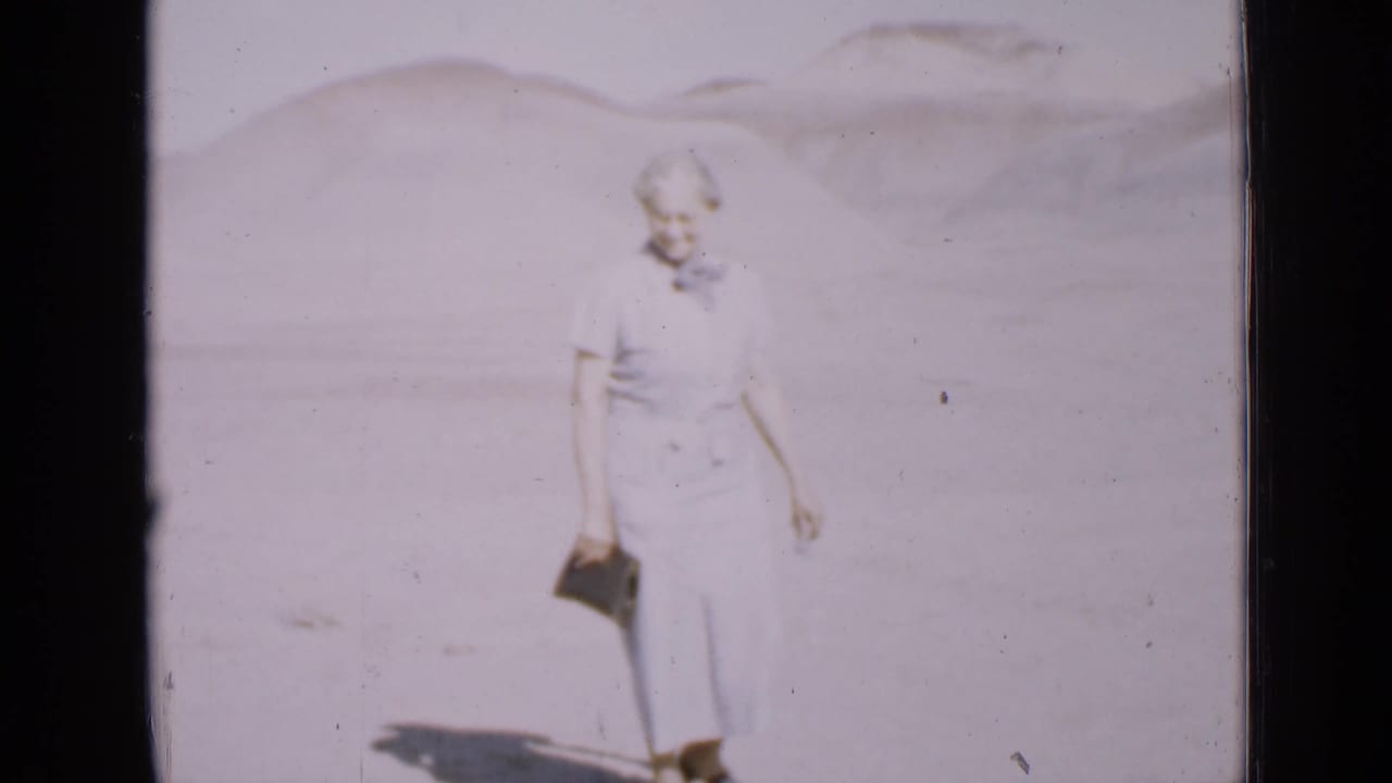 The woman remains in the Utah desert scene, holding her clutch as the faded 8mm frame shows pale sand and low hills.