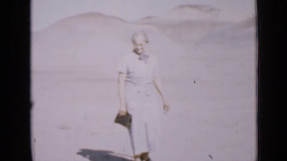 The woman remains in the Utah desert scene, holding her clutch as the faded 8mm frame shows pale sand and low hills.
