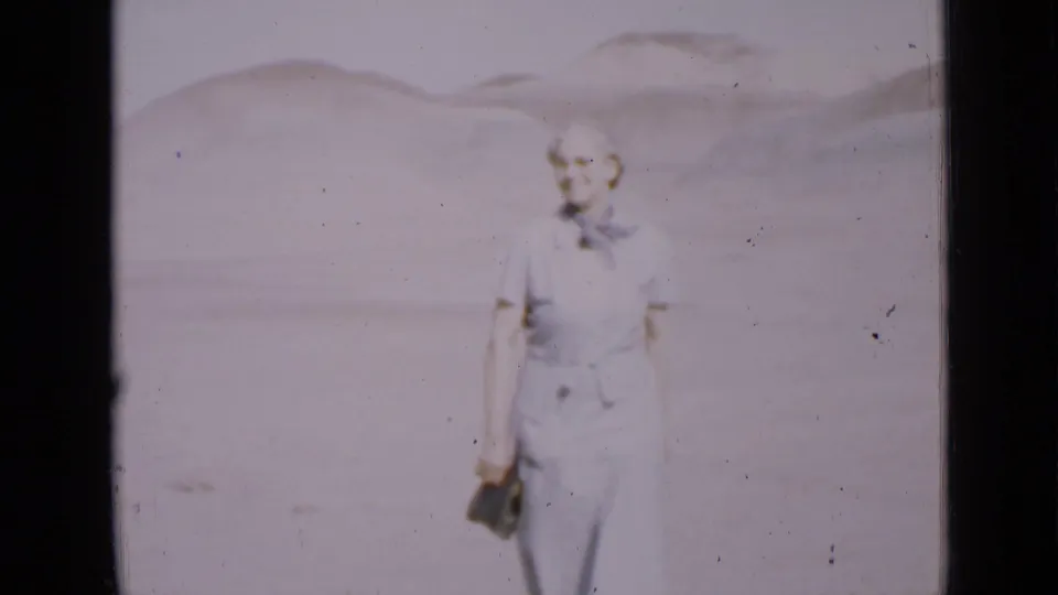 A closer 8mm view shows the older woman in a plain blue dress holding a dark clutch against a bright Utah desert backdrop.