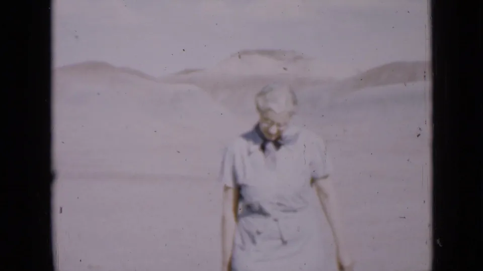 The woman walks slowly across the Utah desert frame, looking down as the sunlit 8mm image fades toward pale blues and whites.