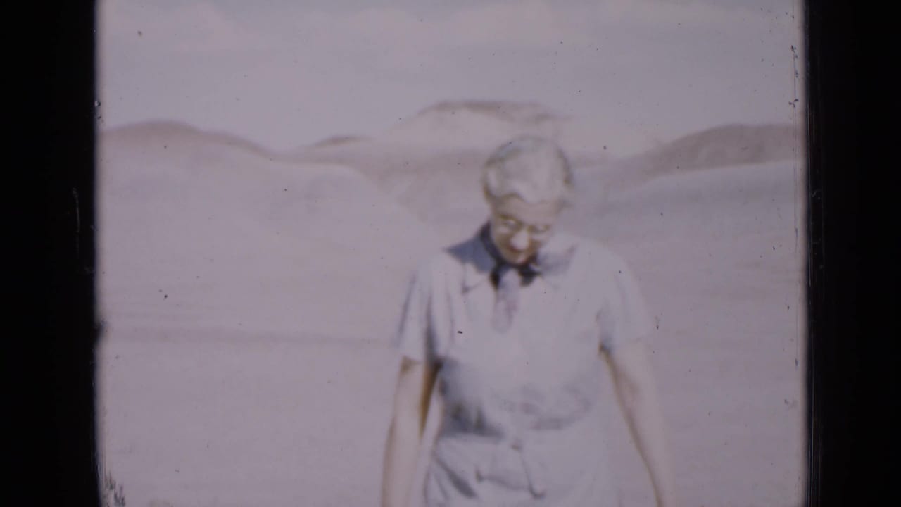 Older woman with short white hair walking on pale sand with dunes behind her.