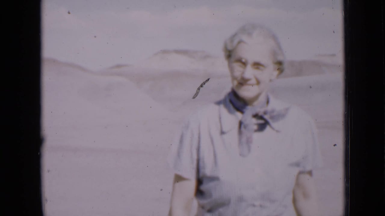 Older woman wearing glasses and a dark neck scarf smiles toward the camera.