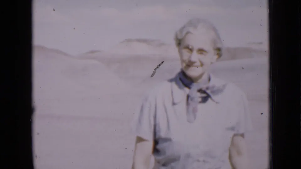 Older woman with glasses smiles toward the camera in a bright Utah desert setting, with dunes behind her.