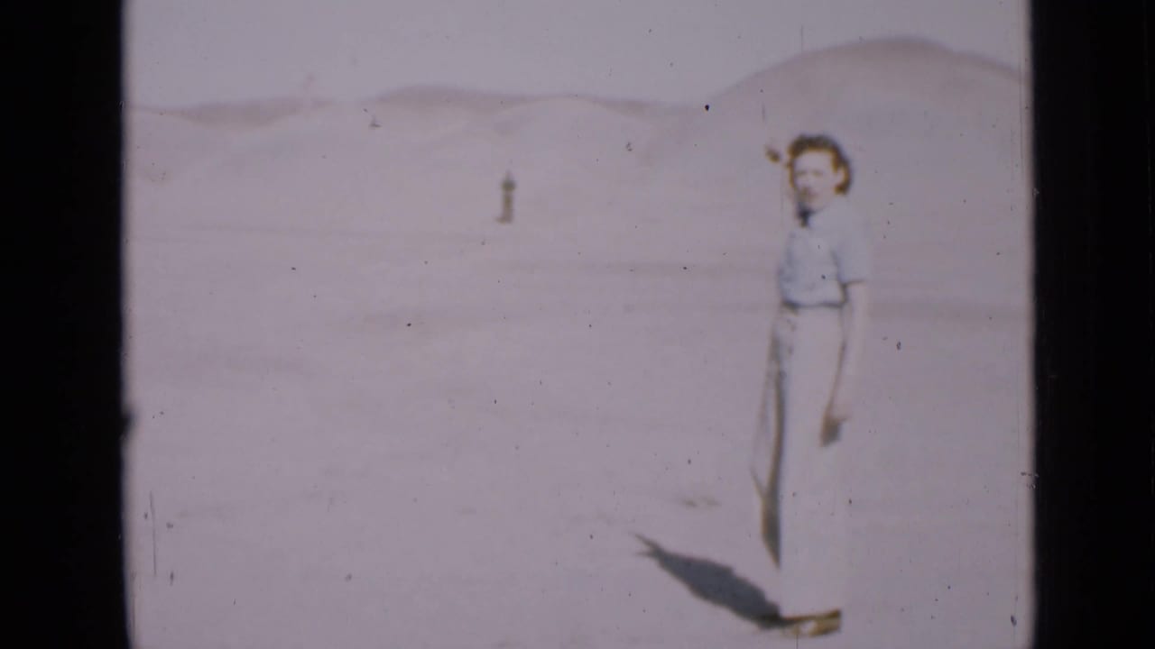 Woman in light clothing stands on pale desert sand with a distant figure behind her.