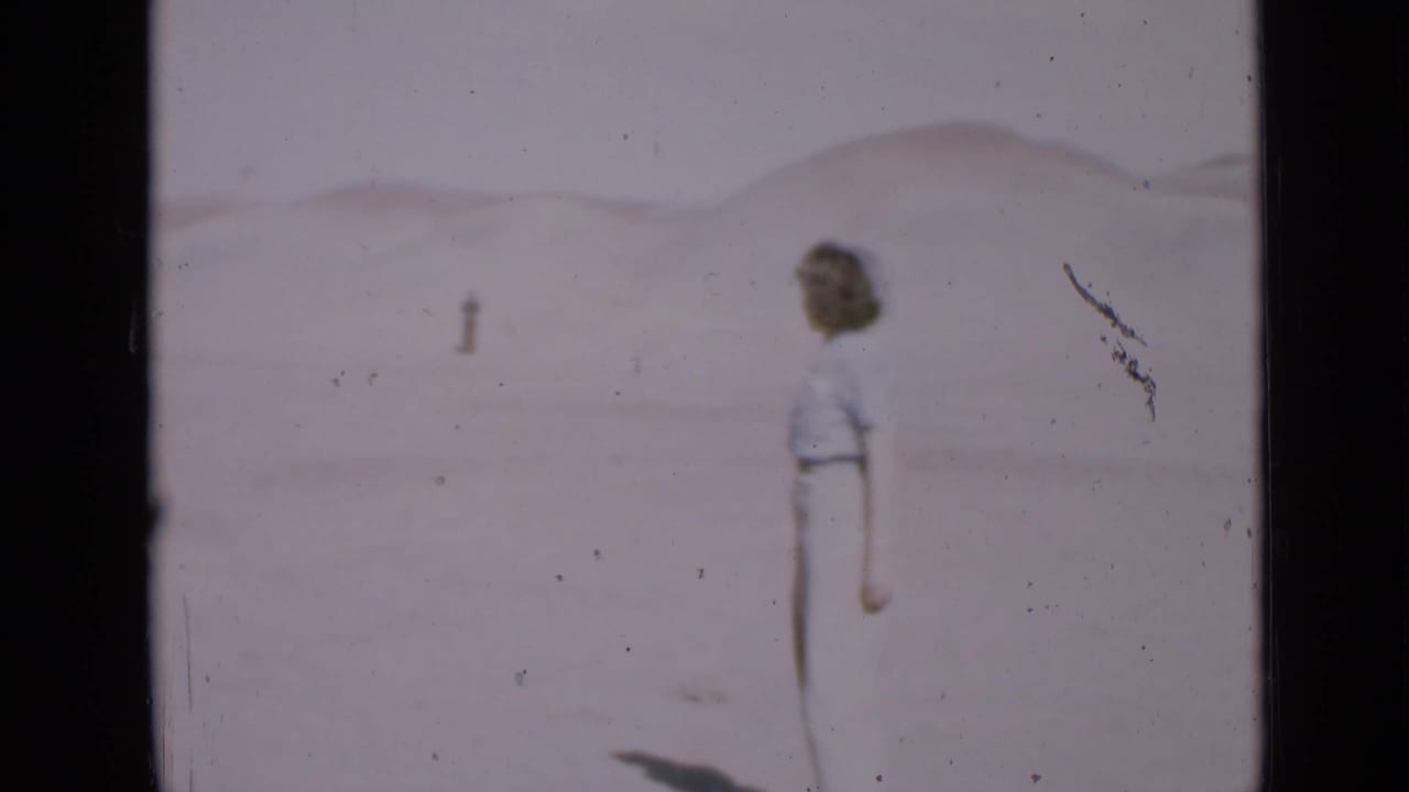 The woman turns away from the camera in the Utah desert, looking toward a small distant figure across the sand.