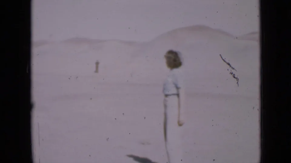 The woman turns away from the camera in the Utah desert, looking toward a small distant figure across the sand.
