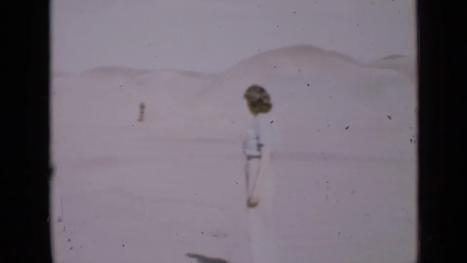 A side view shows the woman standing on pale Utah desert sand, with low dunes and a distant person visible beyond her.