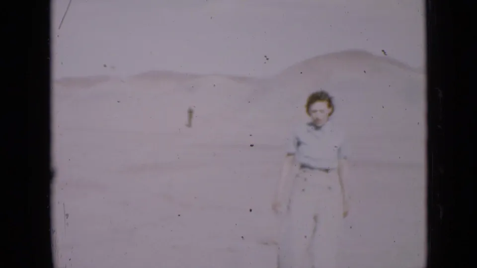 The woman looks back toward the camera in the Utah desert, her shadow falling across the sand while a distant figure remains behind her.