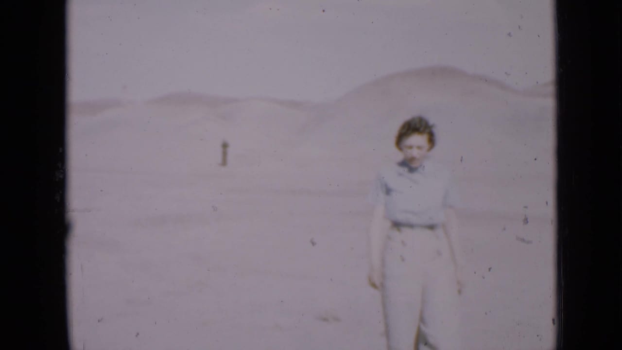 Woman standing in a pale desert landscape with low dunes behind her.