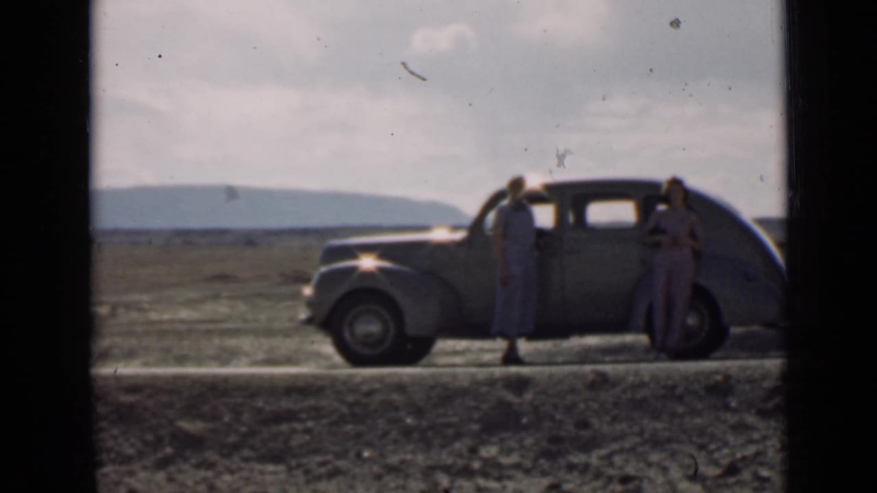 Two women stand beside a rounded vintage car on a dirt road in open country.