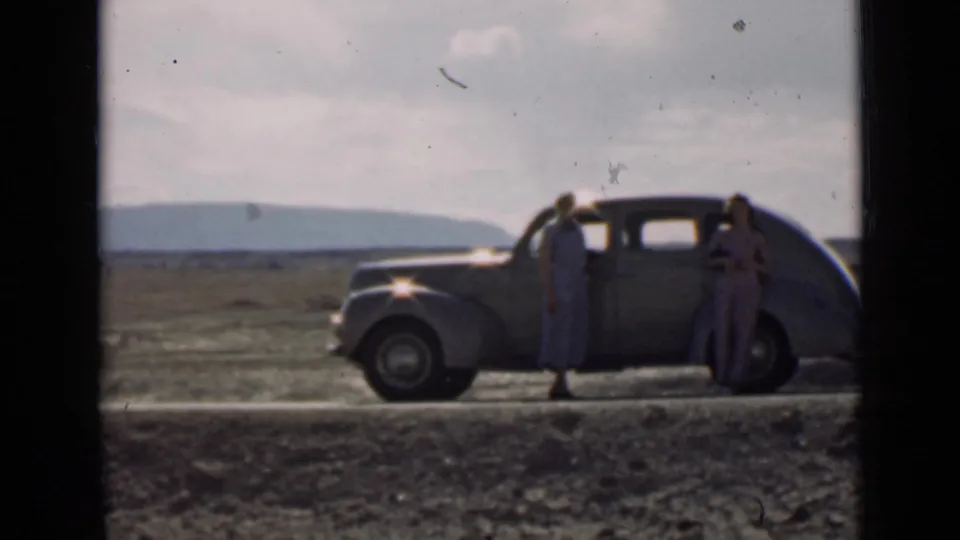 Two women in period dresses stand by a 1930s-style car on a Utah dirt road, with low hills in the distance.