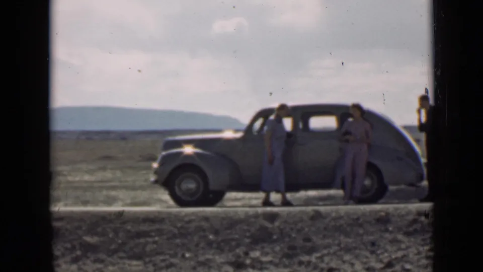 A rounded 1930s-style car sits on a rural Utah road as two women stand beside it and a man appears at the edge of frame.