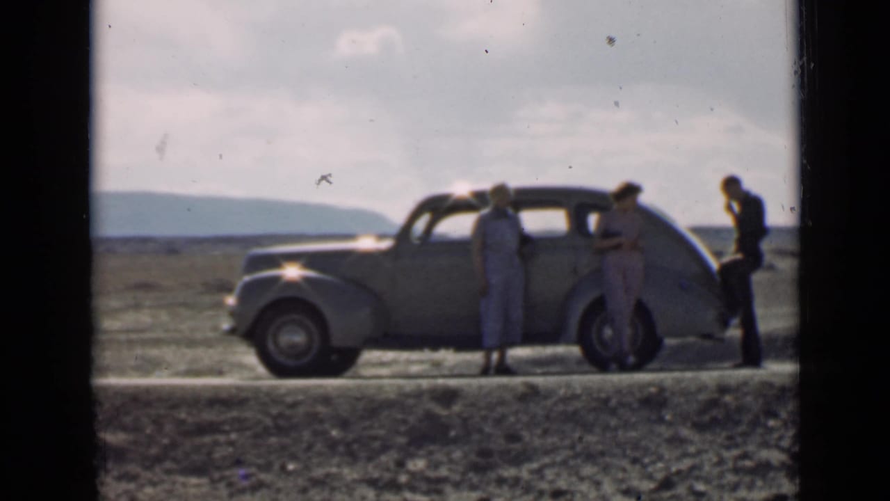 In 1939 Utah home-movie footage, three adults stand near a parked vintage sedan with distant hills beyond the road.