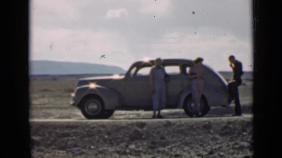 In 1939 Utah home-movie footage, three adults stand near a parked vintage sedan with distant hills beyond the road.