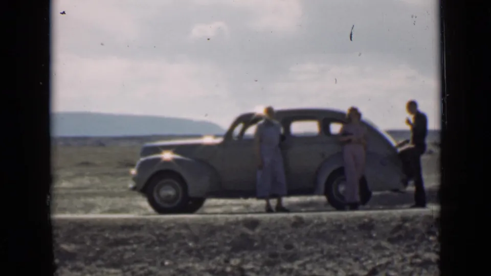 Three adults remain beside a rounded vintage sedan on a Utah roadside, with an open horizon and low hills in the background.