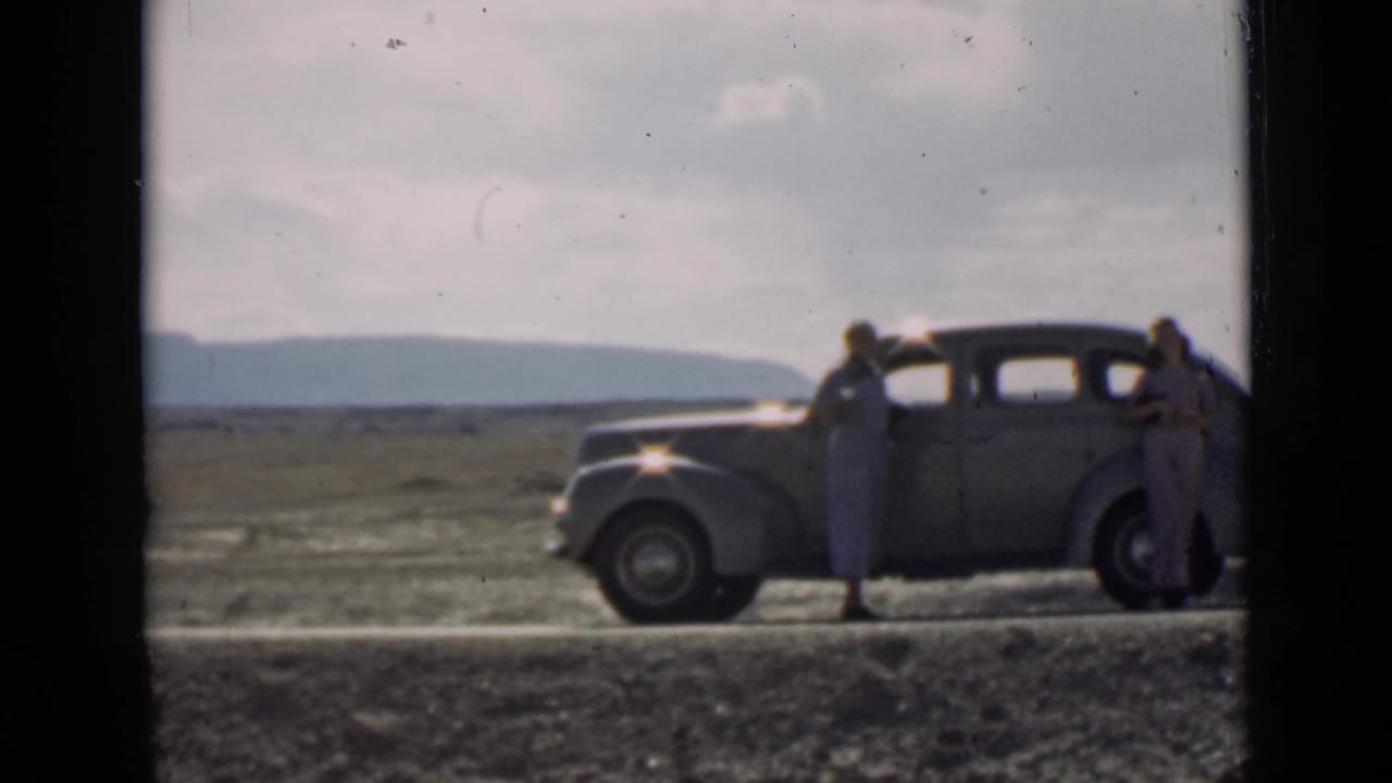 Two women stand beside an older sedan on a remote road with mountains in the distance.