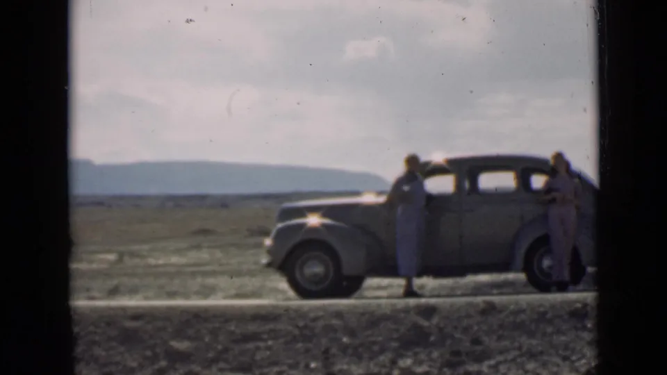 In 1939 Utah home-movie footage, two women stand by a rounded sedan on an open road against a broad arid landscape.