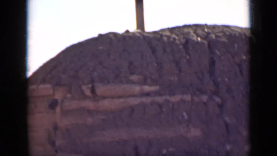The final frame holds on the mud structure's rounded roofline and projecting wooden beams beneath a washed bright sky.