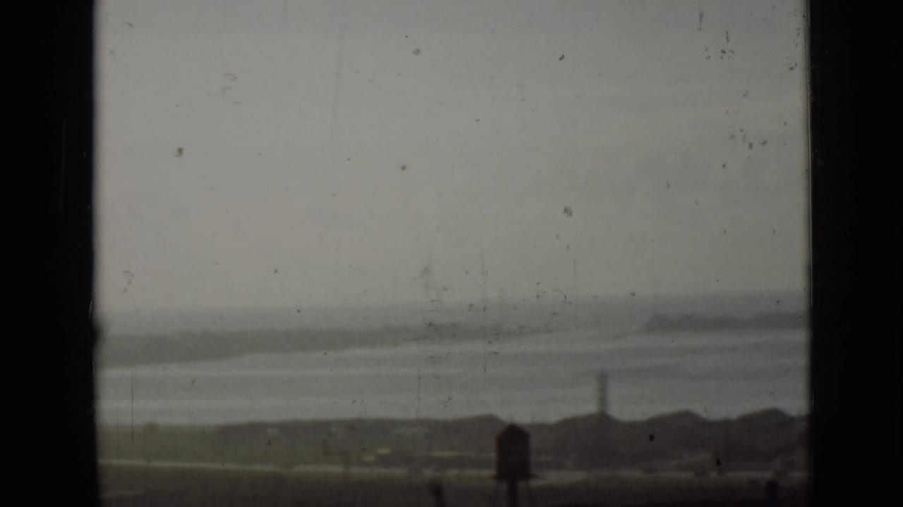 Hazy shoreline with water, low buildings, and a tower beyond dark frame edges.