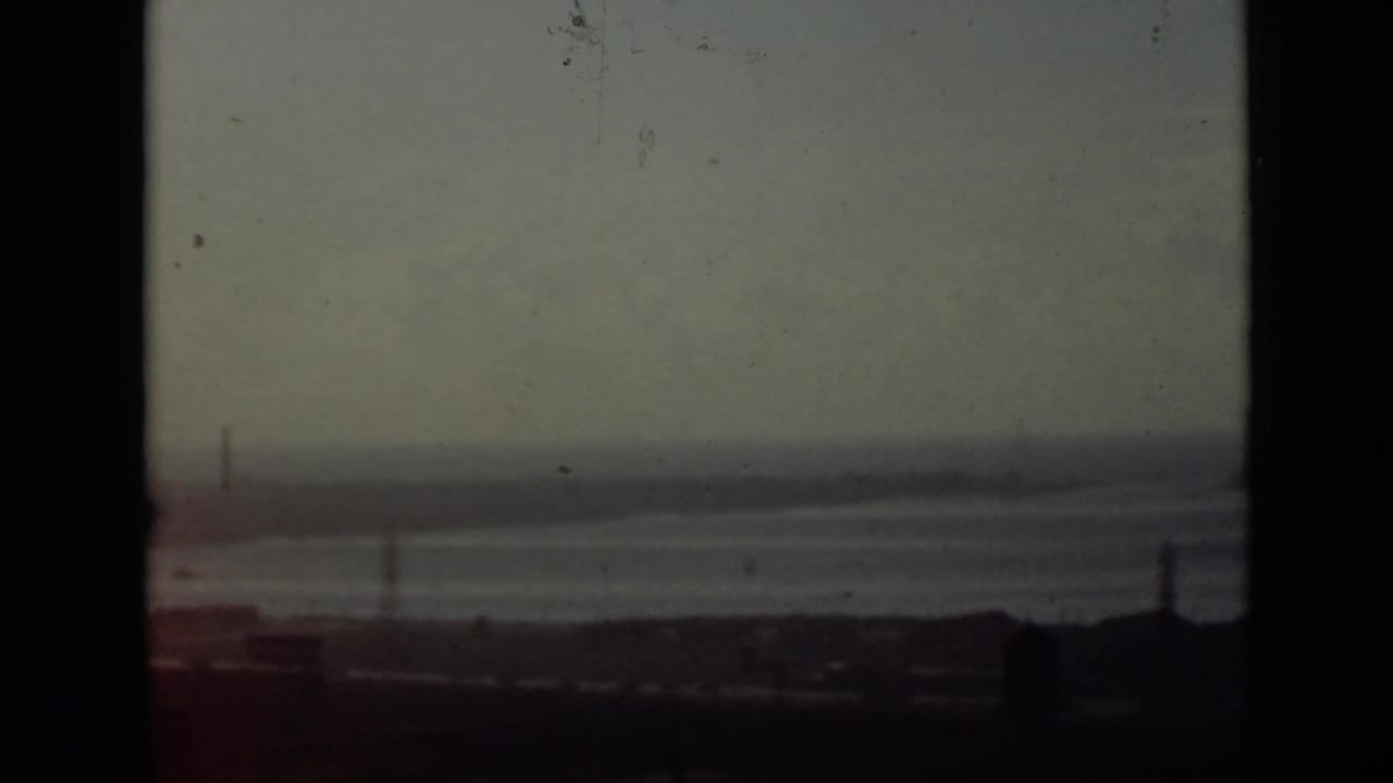 Hazy waterfront seen from above with dark foreground, calm water, distant pylons, and film frame edges.