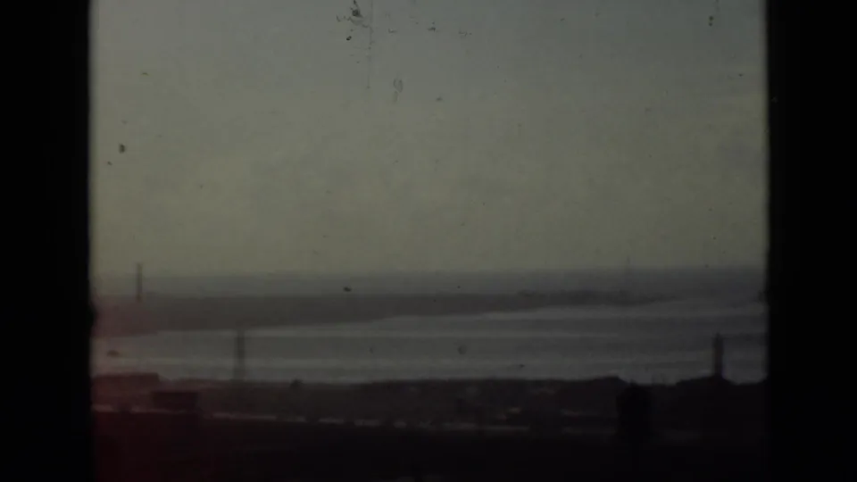 1940 Ontario 8mm frame showing a hazy elevated view across calm water, with a dark shoreline foreground and faint pylons on the horizon.