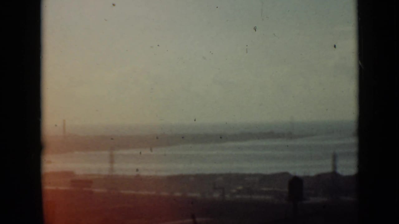Soft waterfront view with calm water, distant shoreline, pylons, dark foreground, and warm film flare.
