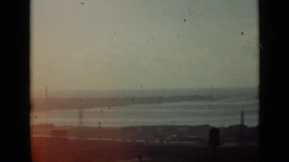 1940 Ontario home-movie frame with calm water beyond a dark foreground, faint pylons, low buildings, and reddish light leak near the lower edge.