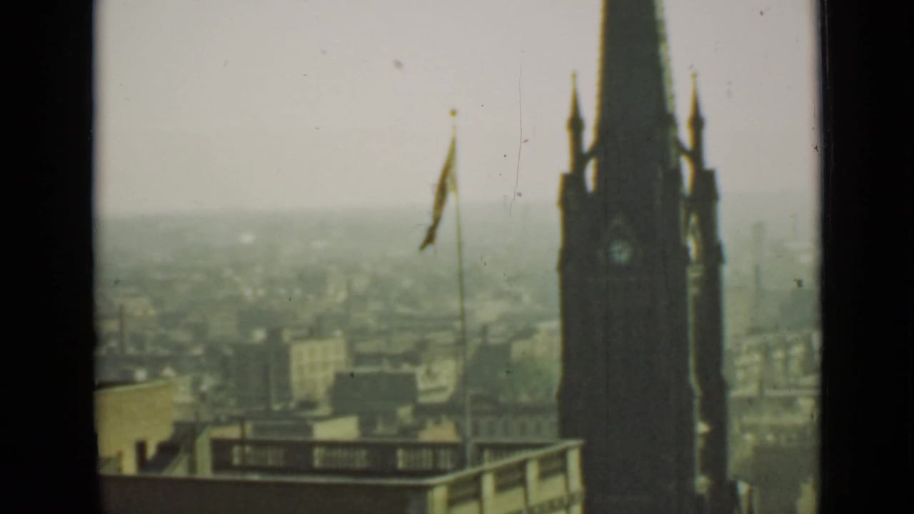 Soft 8mm view of a clock tower, flagpole, foreground roof, and hazy city skyline.