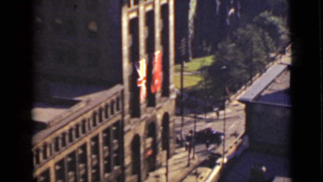 Brick building facade with flags, sidewalk, trees, and a dark car on the street.