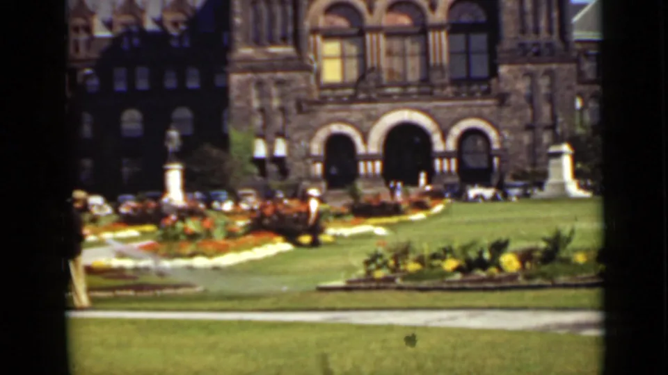 1940 Ontario home-movie frame showing flowerbeds, grass, a statue, and arched stone architecture in bright daylight.