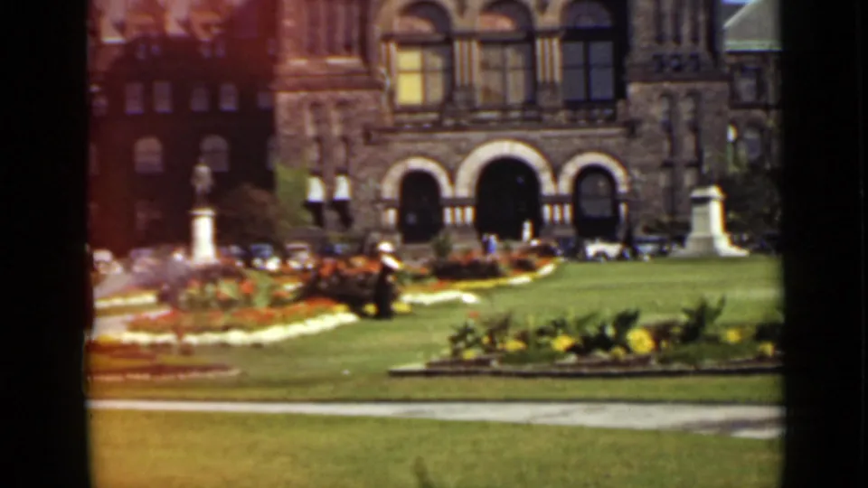 1940 Ontario 8mm frame with visible hose spray over flowerbeds beside a lawn and ornate stone building entrance.