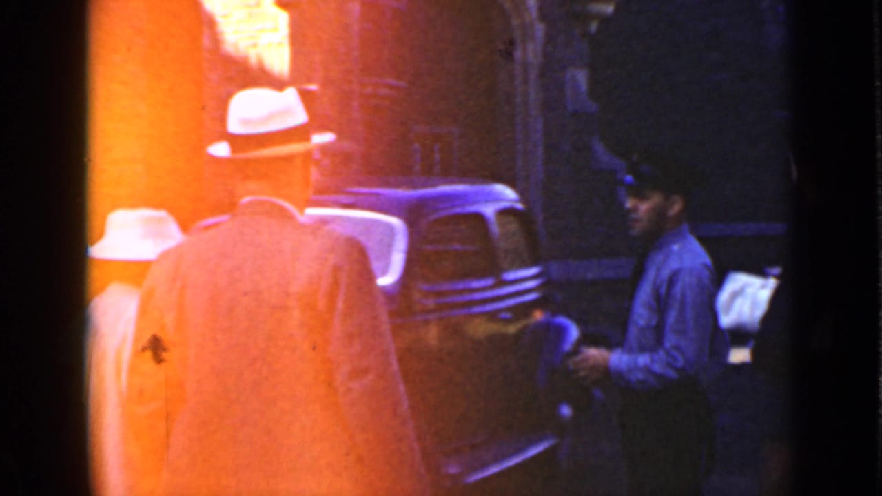 Pedestrians in hats stand near a docked vehicle or vessel area with heavy orange film flare.