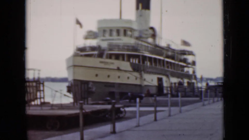 Cream and dark steamship remains moored at an Ontario dock in 1940, framed with railings, posts, flags, and water beyond.