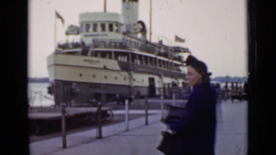 1940 Ontario dock scene with a woman in a dark coat and hat crossing the foreground as a steamship sits moored behind her.