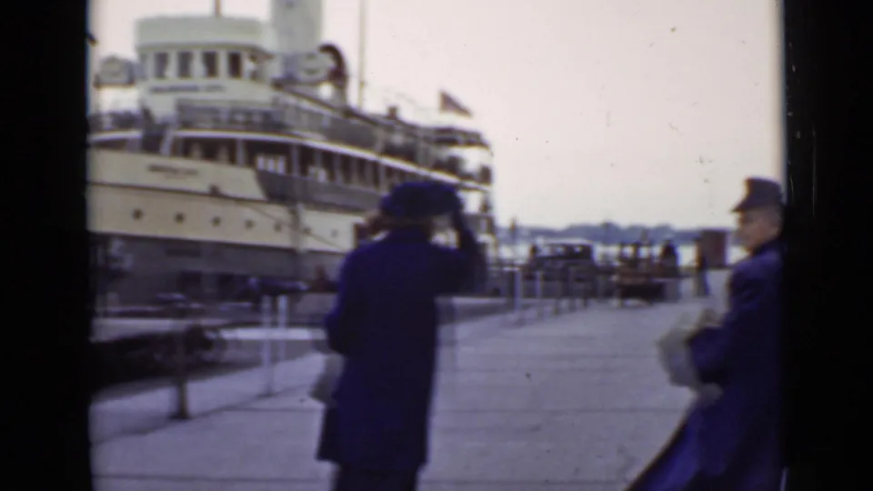Two women in coats and hats move along an Ontario dock in 1940, with the moored passenger steamship and railings behind them.