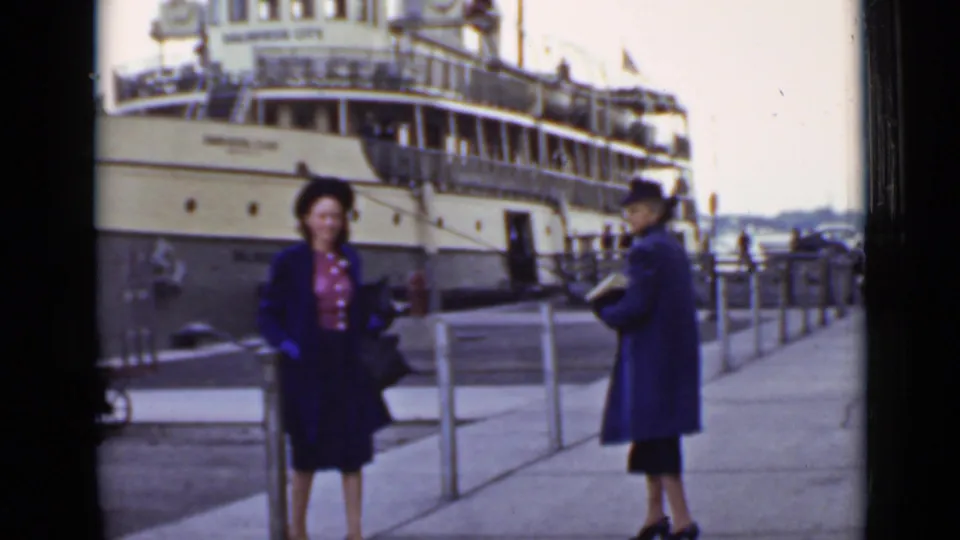 Two women in period coats pose on an Ontario dock in front of a large passenger ship, with railings and water behind them.