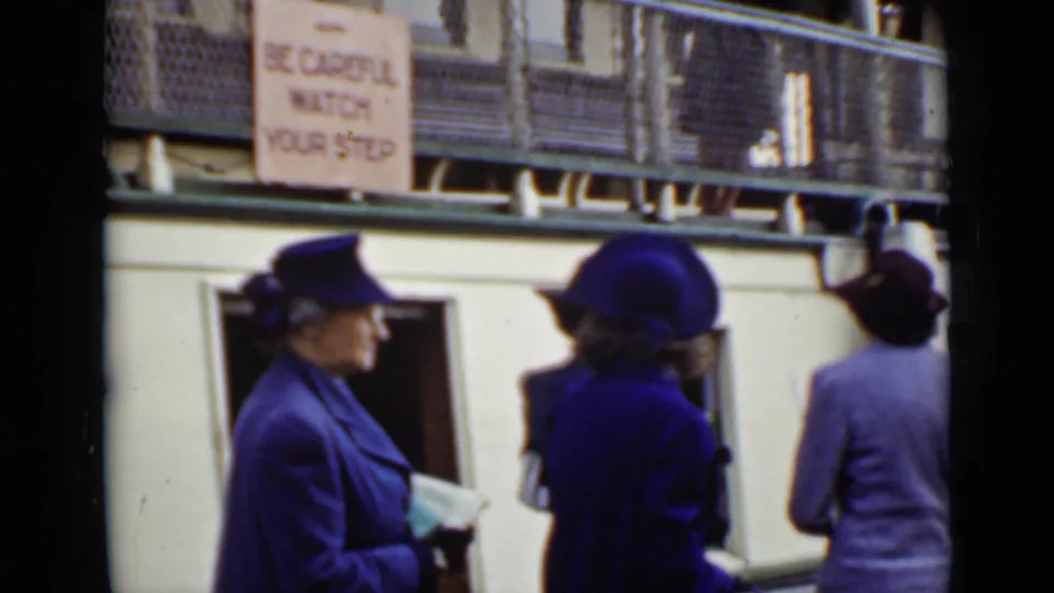 Women in period coats and hats wait beside a boat or dock area in Ontario, with a visible caution sign reading in part, "watch your step."