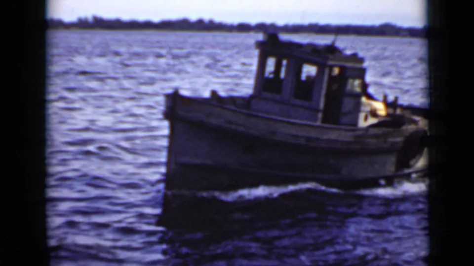 1940 Ontario home-movie frame of a small cabin boat crossing the water, with white foam at the bow and trees beyond.
