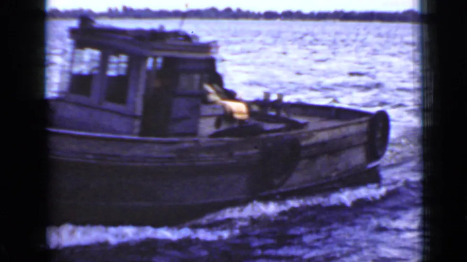 Small cabin boat seen closer in 1940 Ontario 8mm footage, with bright water, low shoreline, and possible occupants obscured inside.