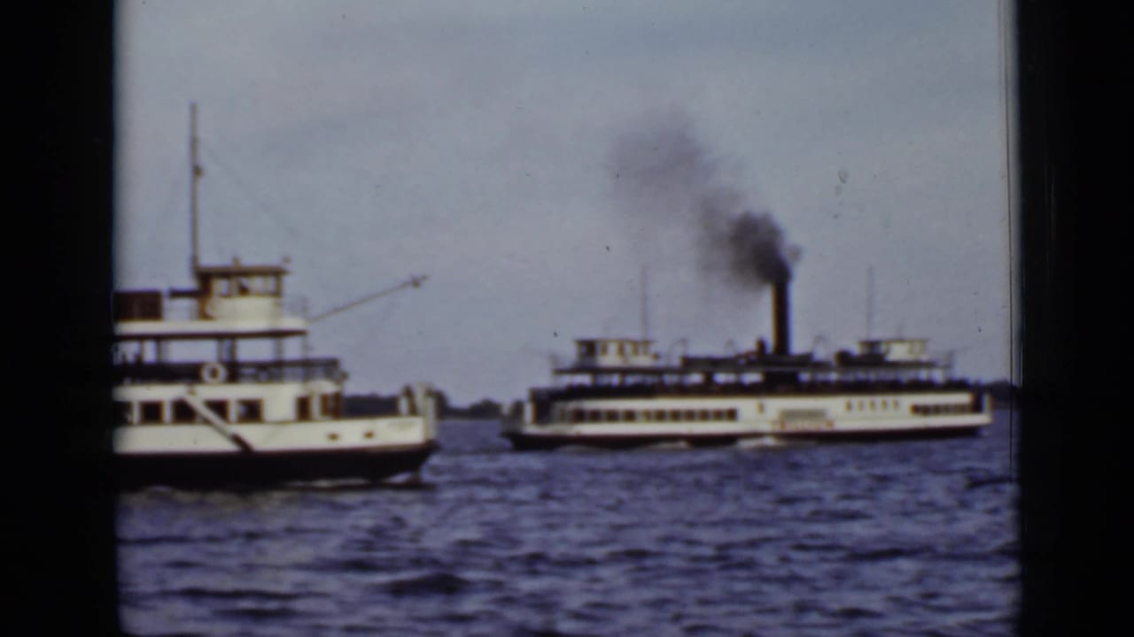 Two ferries pass on open water, with smoke rising from the larger vessel's stack.