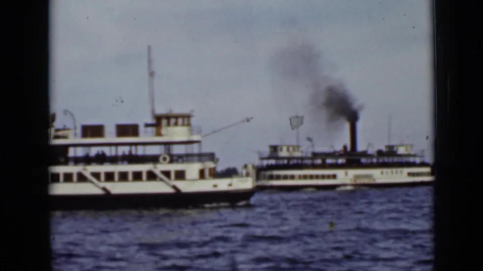 A white multi-deck ferry moves across the foreground on an Ontario lake, with another smoking vessel behind it.