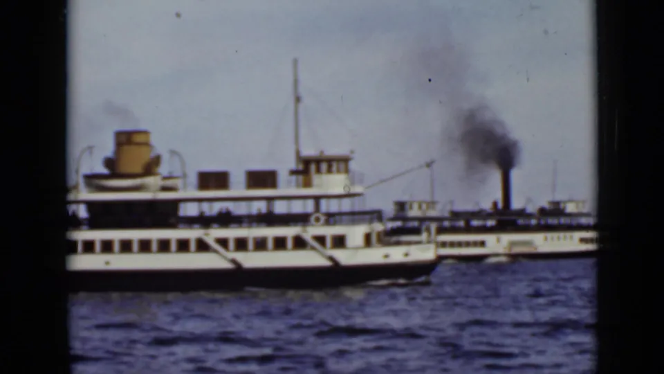 Closer 8mm view of a white ferry's side and decks, with a second smoke-producing ferry partly visible in the background.