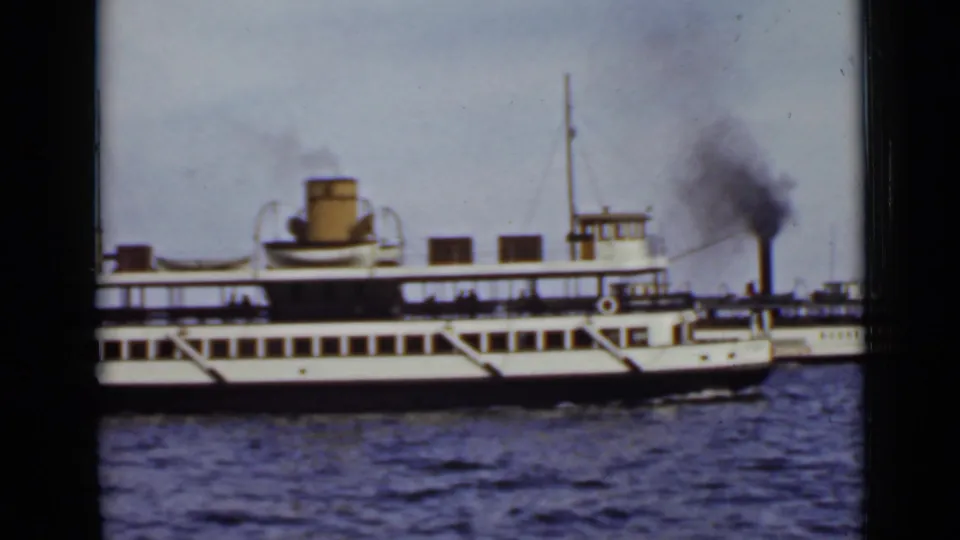 Side view of a white lake ferry with a yellow funnel, while smoke rises from a second vessel near the right edge.