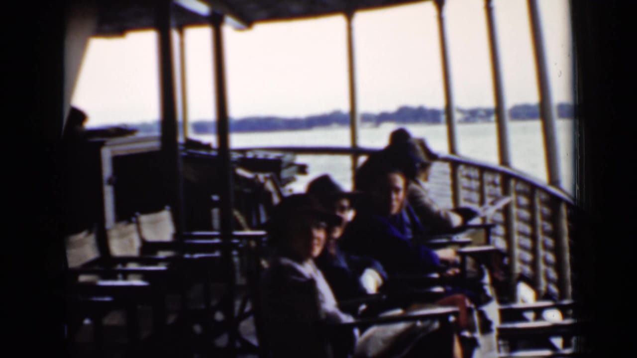 Passengers sit on a covered boat deck beside windows overlooking a lake.